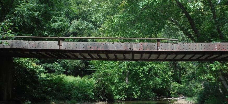 A rusty brown bridge over a creek surrounded by green trees.